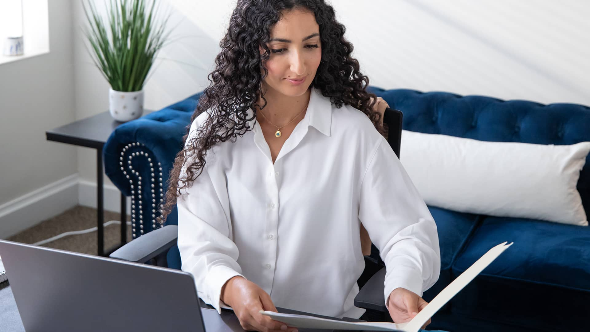 Fatma Ouled Salem, who earned her degree from SNHU, sitting in an office and looking at an open folder she's holding with a blue couch and potted plant on a small table in the background.