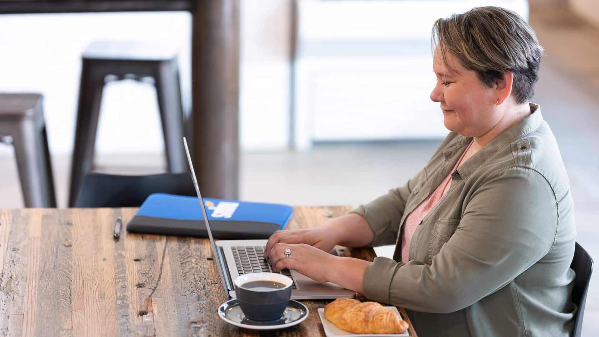 Alaine Garcia, who earned her degree from SNHU in 2020, sitting at a wooden table typing on her laptop with  an SNHU notebook, a cup of coffee and a croissant on the table next to her.