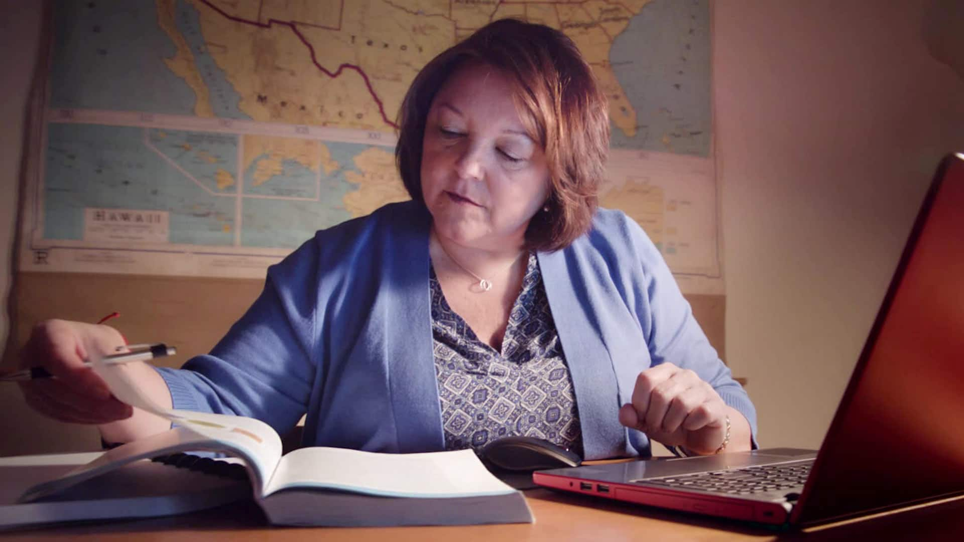 Bonnie Fecowicz, who earned her degree from SNHU in 2016, sitting at a desk in a dim office, looking at a page in a text book with an open laptop in front of her.