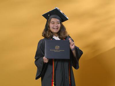 Adrian Henares '24, a Maryland resident who earned her bachelor's in communication, wearing her cap and gown and holding her diploma.