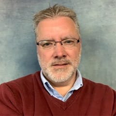 Thomas Jensen, an adjunct marketing faculty member at SNHU wearing a red shirt on a grey background.