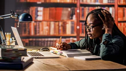 A student sitting at a desk in a library taking notes from a laptop