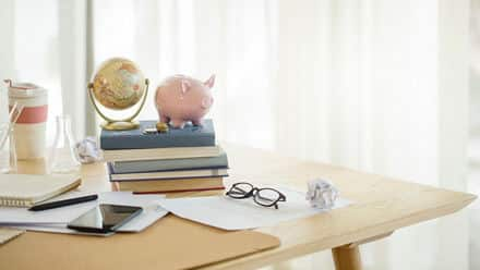 A desk with papers, a phone, and a stack of books with a piggy bank and globe on top