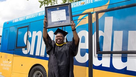 Clad in graduation regalia and standing in front of an SNHU bus, Lorenzo Mateo '19, holds up a framed diploma after earning a BS in Information Technology.