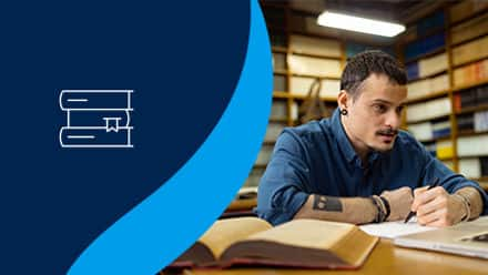 A student sitting in a library working on their english degree with a stack of books to the left on a blue background.