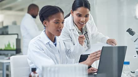 Two women in lab coats working in health science looking at a computer with a co-worker in the lab behind them