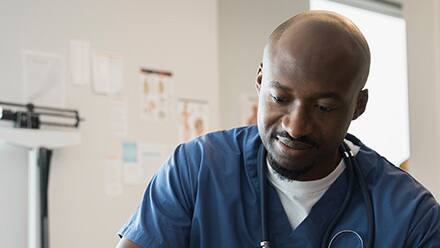 A male nurse wearing blue scrubs and a stethoscope writing in a medical chart.