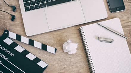 A screenwriters desk with a laptop, movie clapper, headphones a script and crumpled up paper of screenplay drafts.