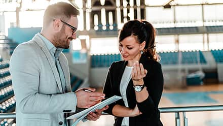 Two sports management professionals in a gym, discussing notes from a notebook.