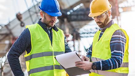 Two engineers wearing hard hats and safety vests consulting a clipboard