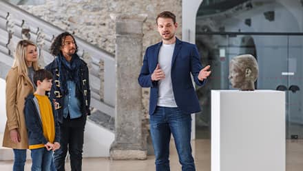 A curator with a master's in history, gesturing to a sculpture of a head during a museum tour.