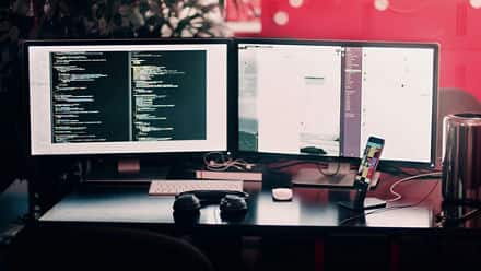 The desk of a web developer with two desktops open on development work along with a pair of headphones and a phone.