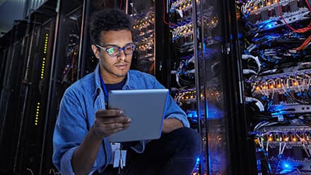 A student exploring what cyber security is, holding a tablet and standing in front of large machines at his internship.