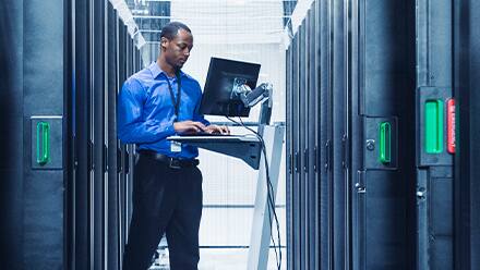 A man uses his cyber security degree while working on a laptop inside a server room.