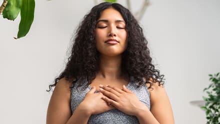 A woman surrounded by plants, meditating with her eyes closed and hands over her chest