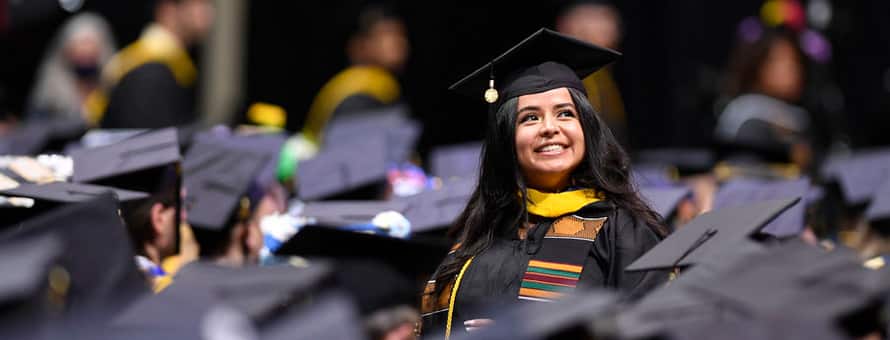 An SNHU graduate in cap and gown, standing among many other sitting graduates.