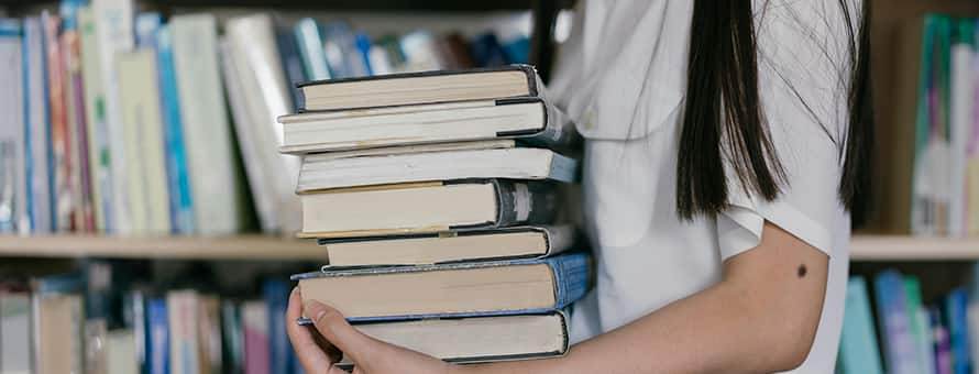 A student holding a stack of books in a library working on academic referencing for their research paper.