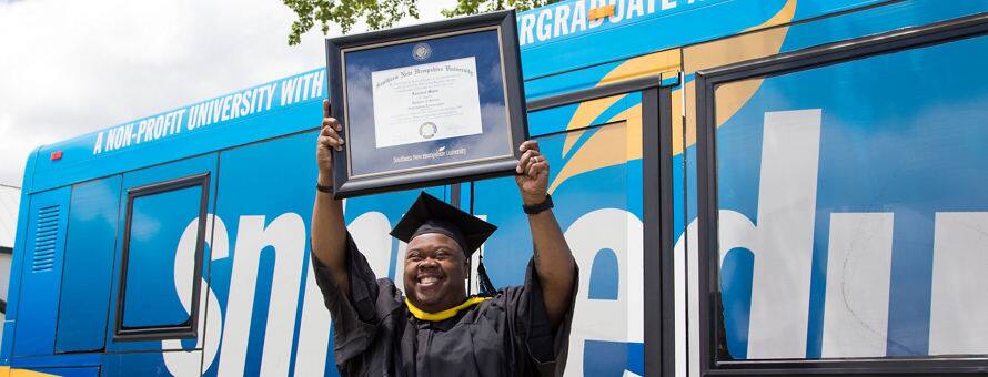 Clad in graduation regalia and standing in front of an SNHU bus, Lorenzo Mateo '19, holds up a framed diploma after earning a BS in Information Technology.