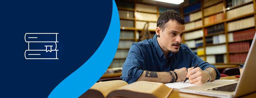 A student sitting in a library working on their english degree with a stack of books to the left on a blue background.