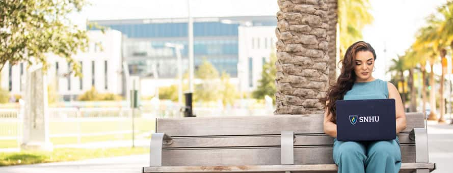 SNHU student sitting on a bench in front of a palm tree typing on her laptop.