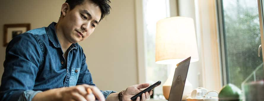 A man holding a cell phone and in front of his laptop as he works remotely.