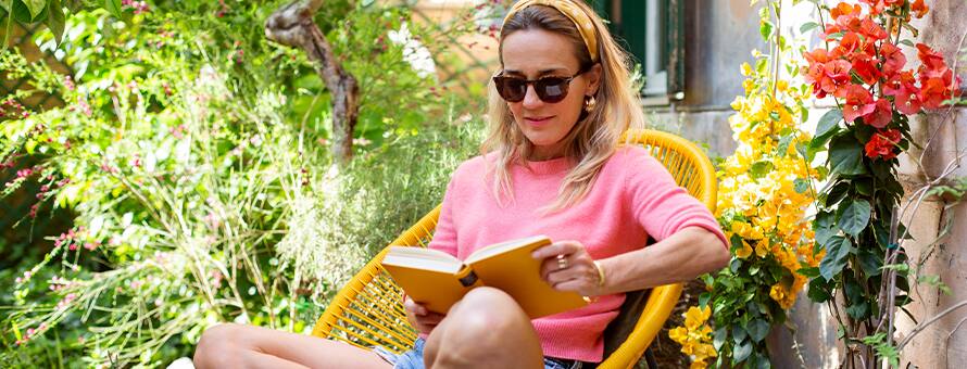 A woman with blonde hair in a pink shirt sitting in a yellow chair reading a book in the summer