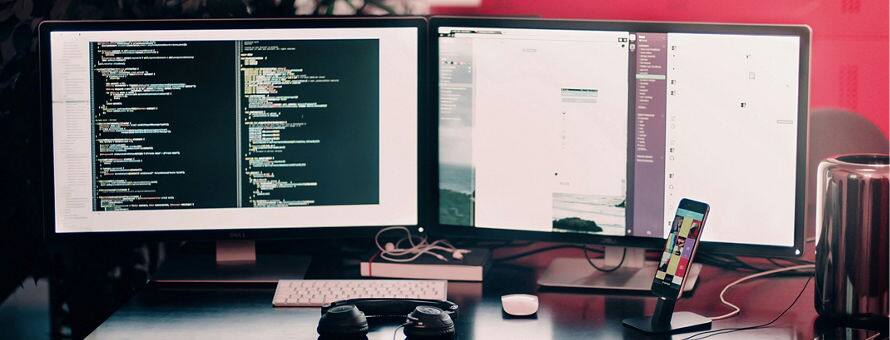 The desk of a web developer with two desktops open on development work along with a pair of headphones and a phone.