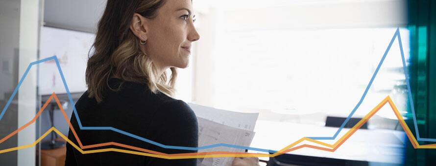 An economist reviewing paperwork at her desk with a blue, orange and yellow line graph overlay.