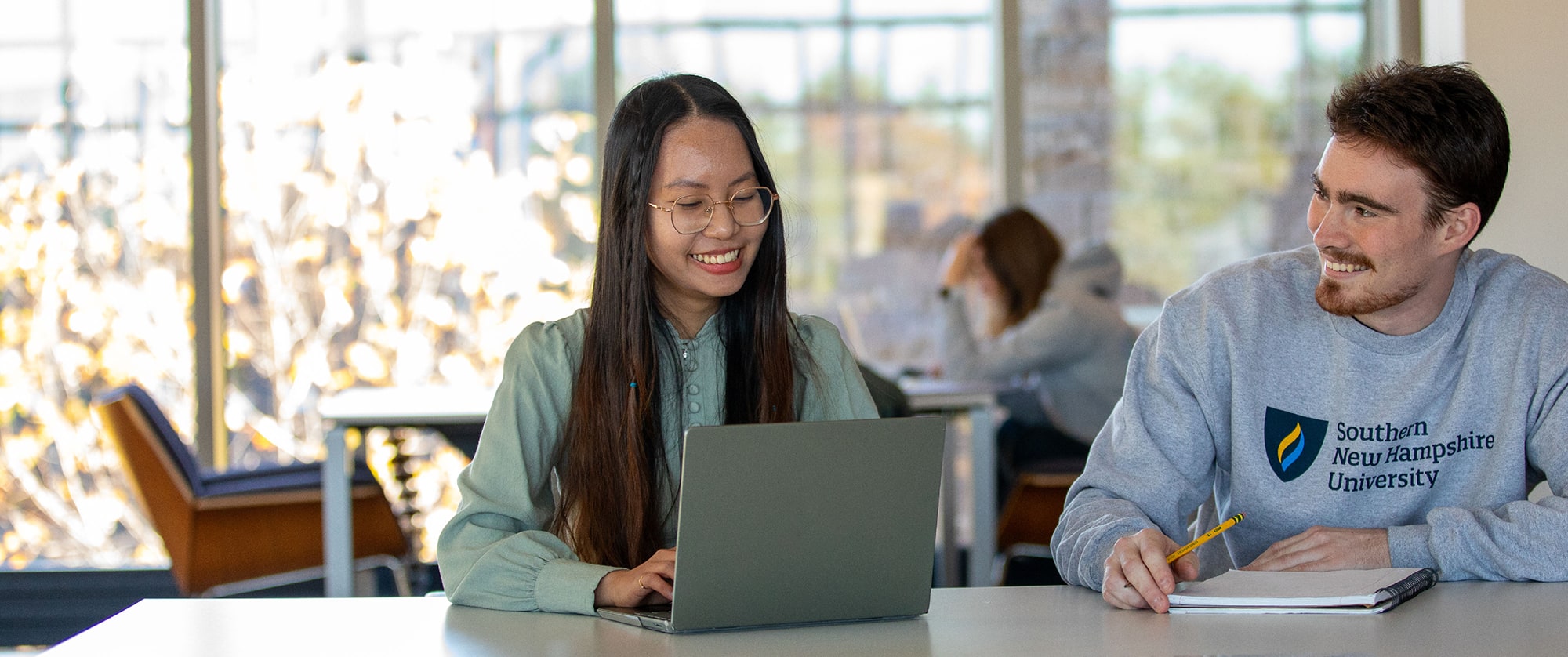 Two students collaborating at a table in the library. One student is using a laptop while the other takes notes, wearing a Southern New Hampshire University sweatshirt.