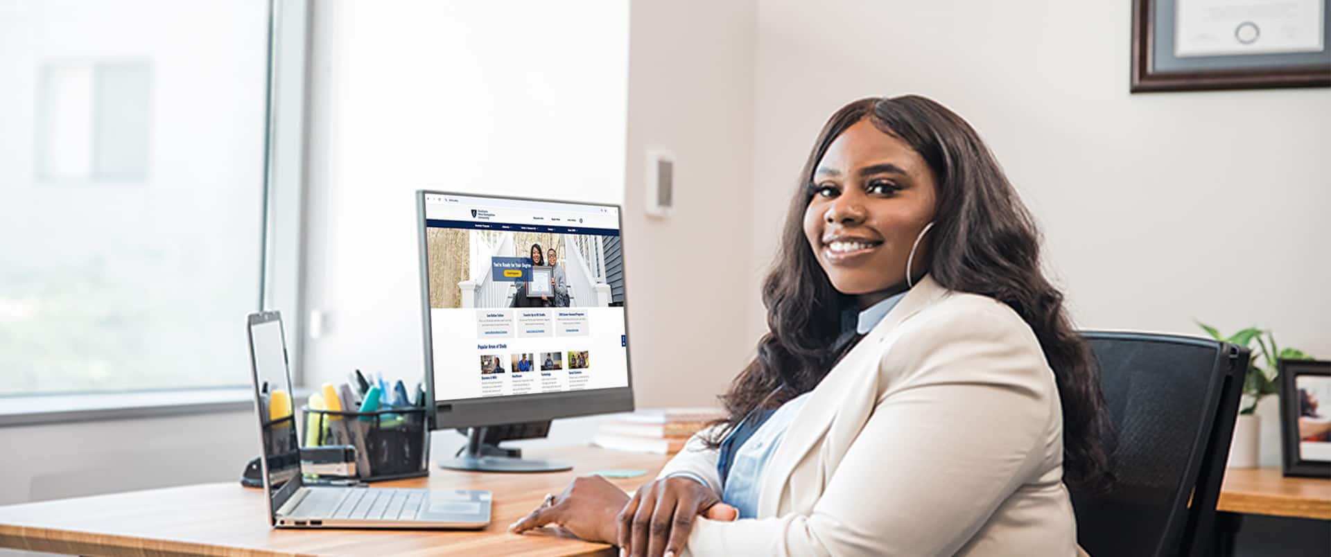 Tanzania Fair, who earned her degree at SNHU, sitting at an office desk with a computer monitor, a laptop and pen case in the background.