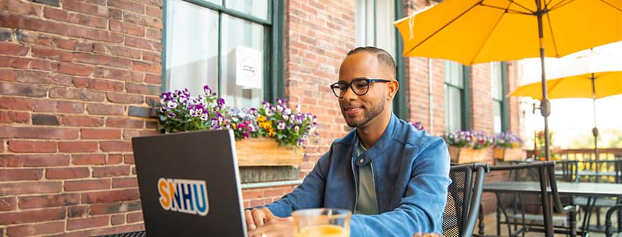 Geremy Laboy Prospere, sitting at an outdoor cafe working on a laptop with an SNHU sticker on the cover.