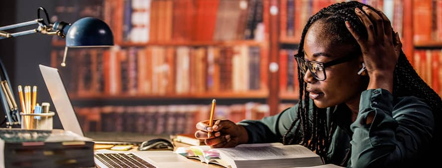 A student sitting at a desk in a library taking notes from a laptop