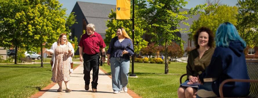 2 students walking on campus with a professor, while 2 other students sit on a bench.