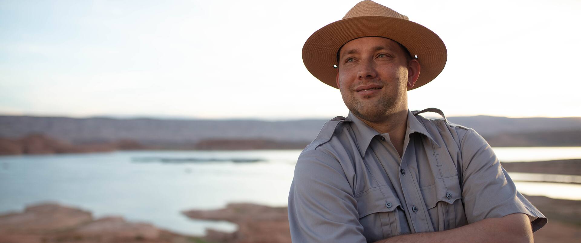 John Roos, who earned his degree in 2018, wearing a park ranger hat and grey buttondown shirt standing with his arms folded. 