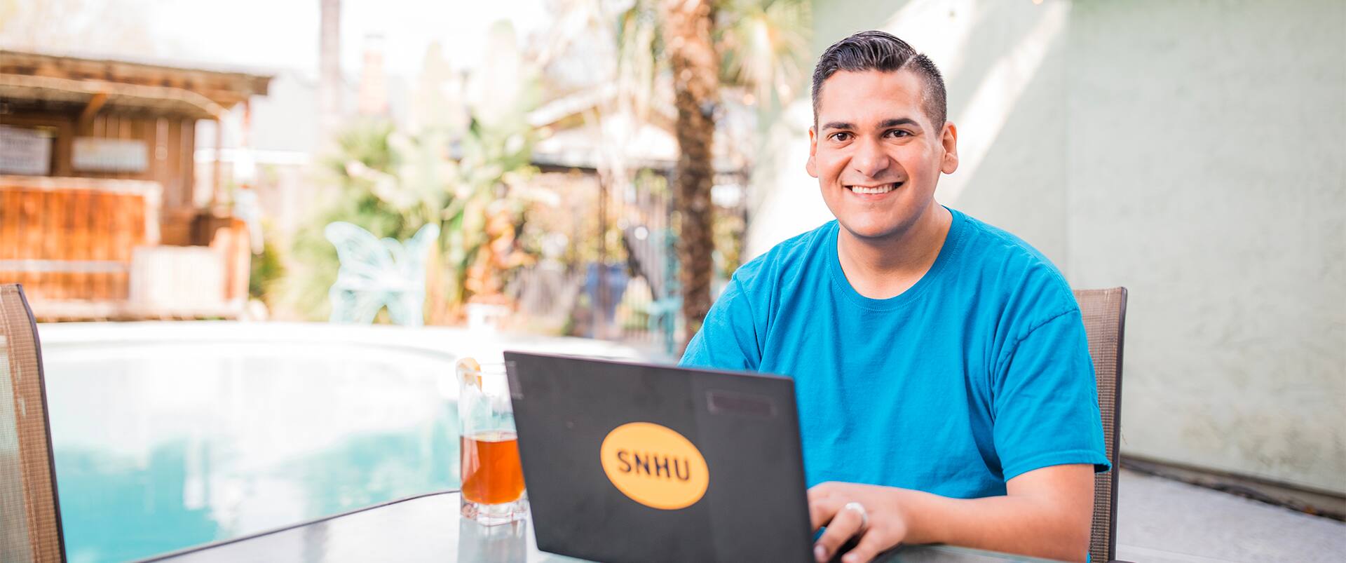 Juan Munoz, who earned his general studies degree in 2022, smiling and working on his laptop beside a pool and with a glass of ice tea with a lemon beside him.