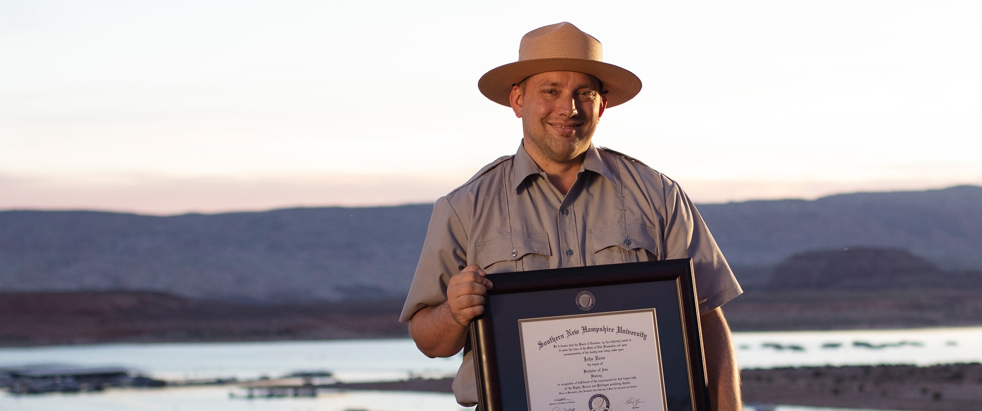 John Roos, who earned his history degree in 2018, wearing his park ranger uniform and hat and holding his framed SNHU degree with a lake and small mountains in the distance behind him. 