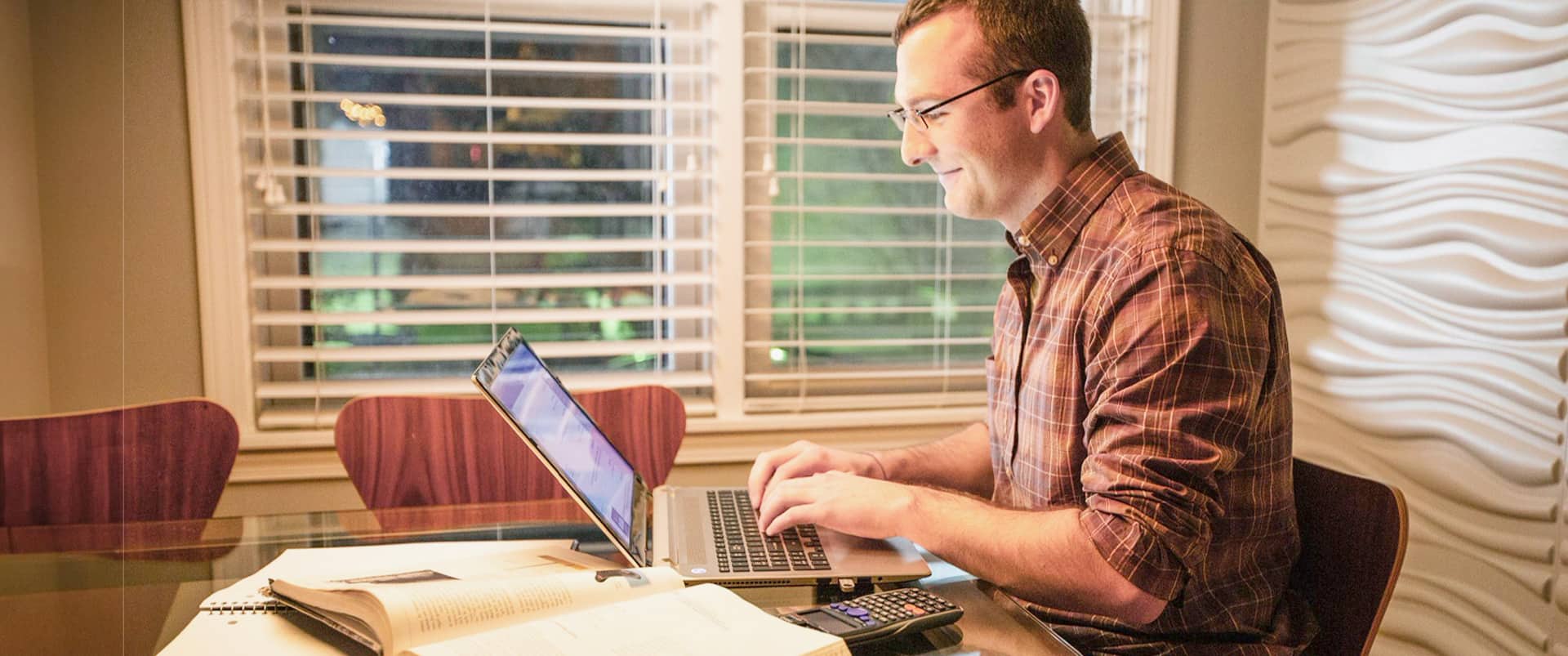 Chris Eldridge, who earned his degree from SNHU in 2015, wearing a plaid shirt, sitting at a glass table, typing on a laptop with a scientific calendar and open notebooks and text books beside him.
