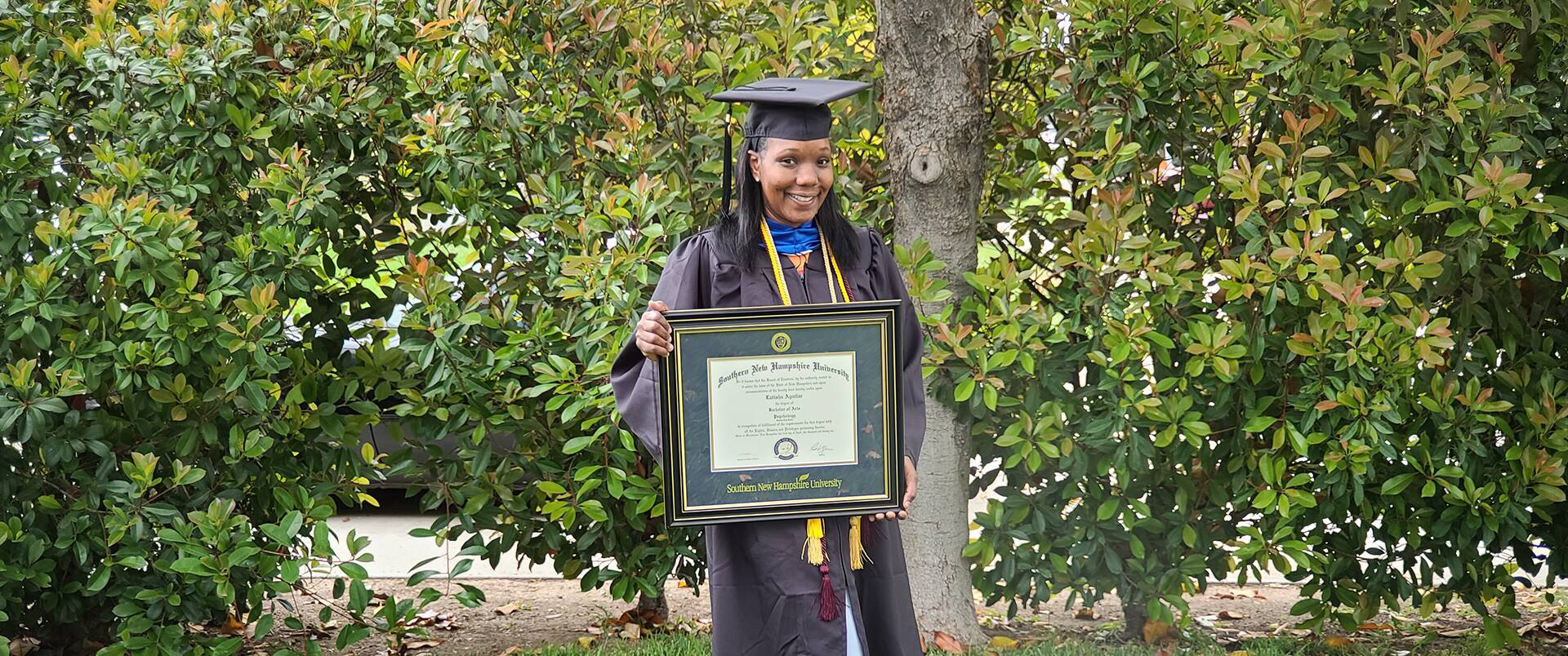 Latisha Aguilar holding her bachelor's in psychology degree diploma while standing in front of a tree.