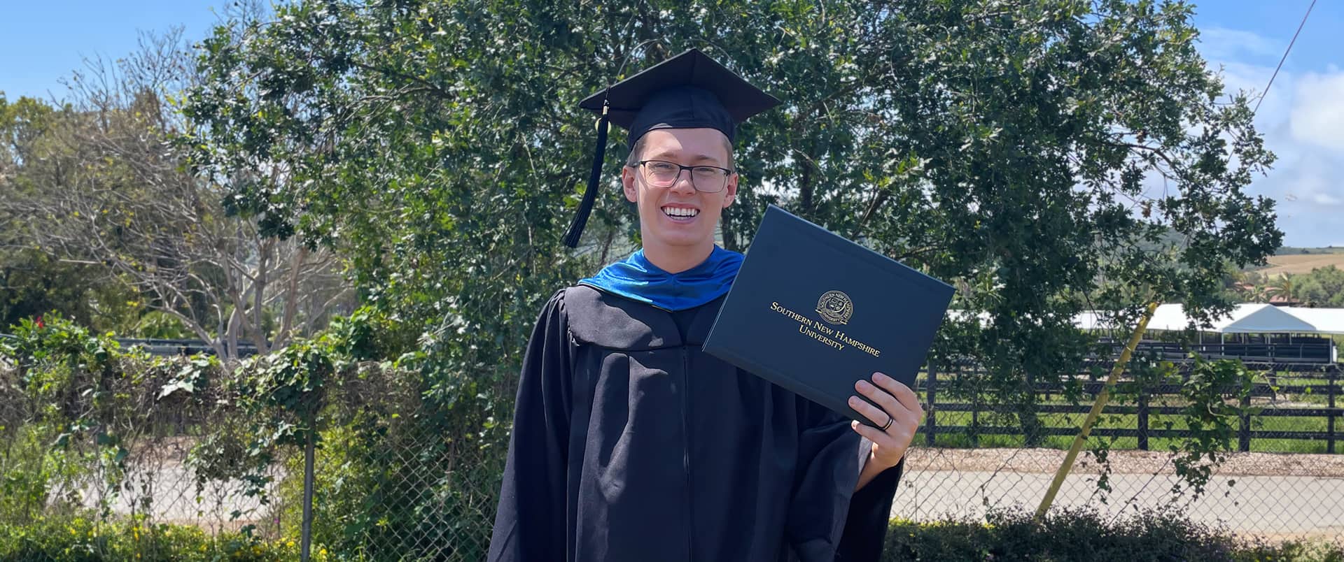 Matthew Daigneau, who earned his degree from SNHU, standing outside in front of a small tree wearing his cap and gown and holding his diploma.
