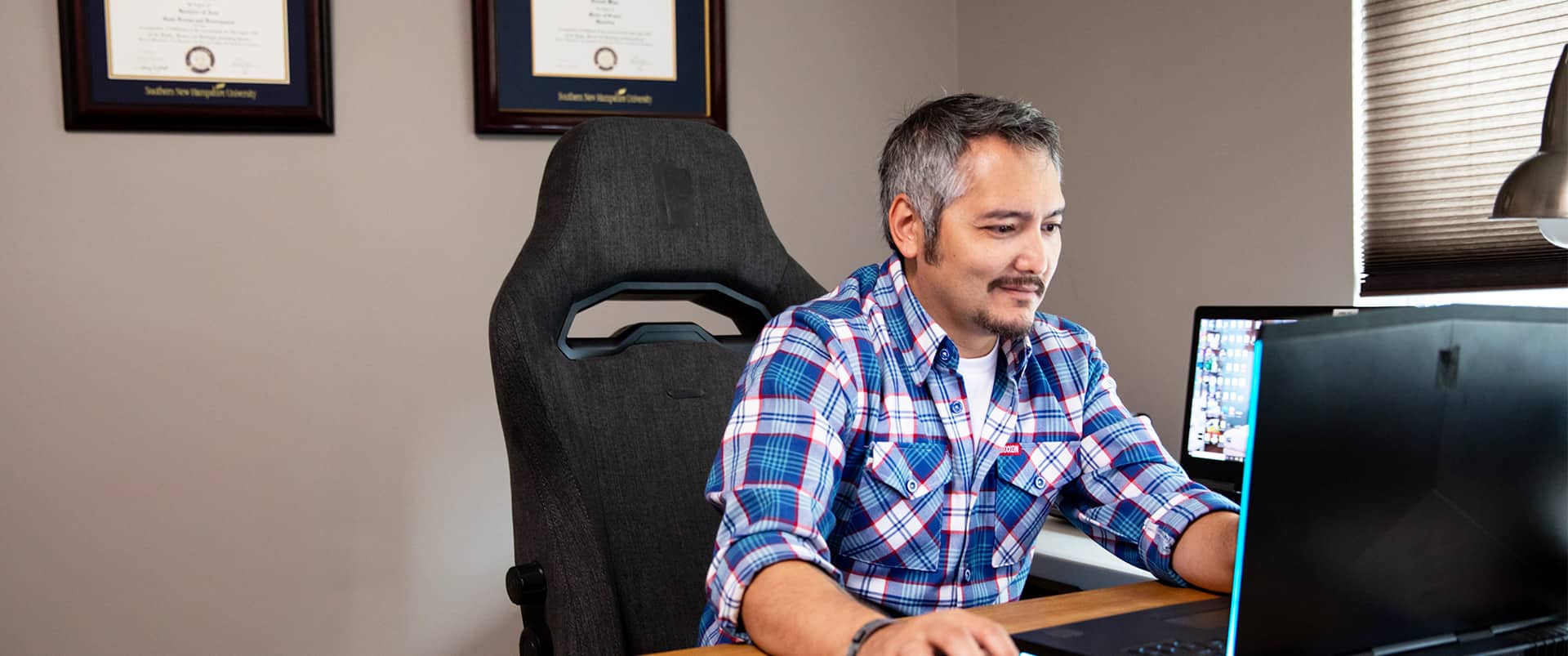 "Derald Wise, who earned degrees from SNHU in 2014 and 2018, sitting at a desk  working on a laptop with two framed SNHU degrees on the wall behind him."