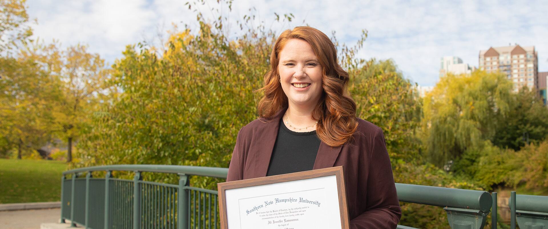 Ali Lamoureux-Rule holding her 2022 BS in Healthcare Administration diploma while standing on a footbridge in a park.