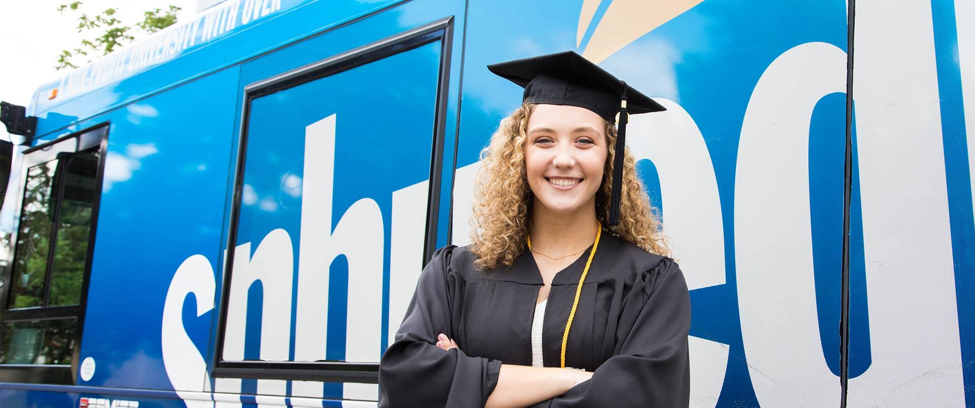 Joann Coffey, who earned her degree from SNHU, wearing her cap and gown and standing in front of the SNHU-branded bus.