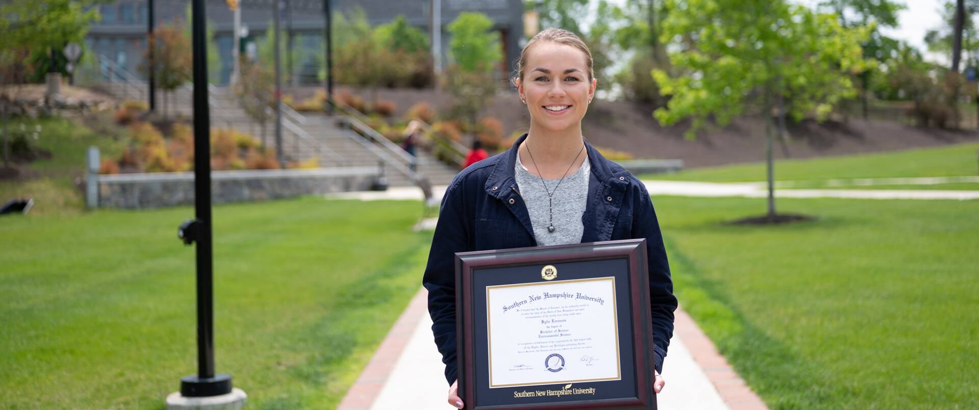 "Kylie Lorenzen, who earned her degree from SNHU in 2019, holding her framed diploma  while standing in the quad on the SNHU campus."