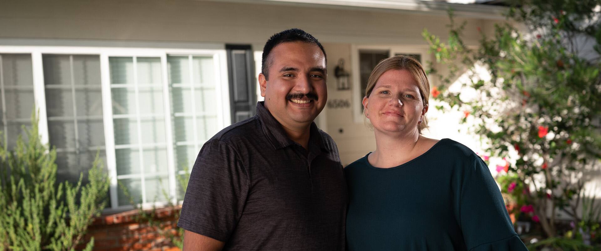 "Shelly & Salvador Villa, a married couple who earned their degrees from SNHU in 2019,  standing next to one another in their front yard with plants and a hite house in the  background."