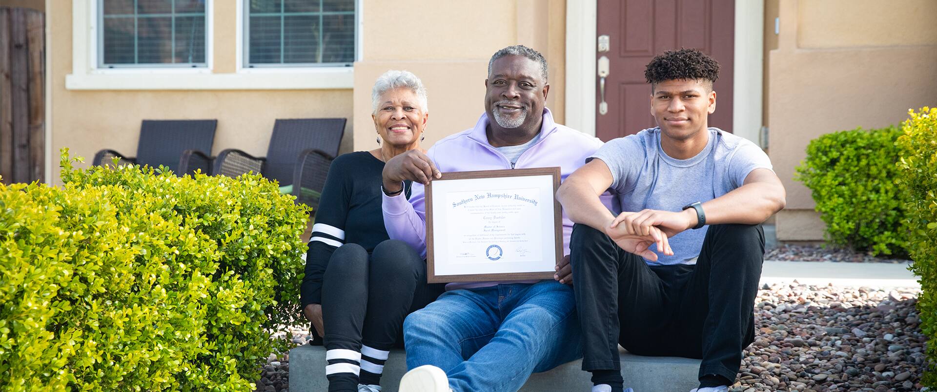 Corey Dantzler, who earned a graduate certificate in athletic administration in 2023, holding his framed SNHU diploma sitting on stone steps in front of his home with a younger man to his left and an older woman with white hair to his right.