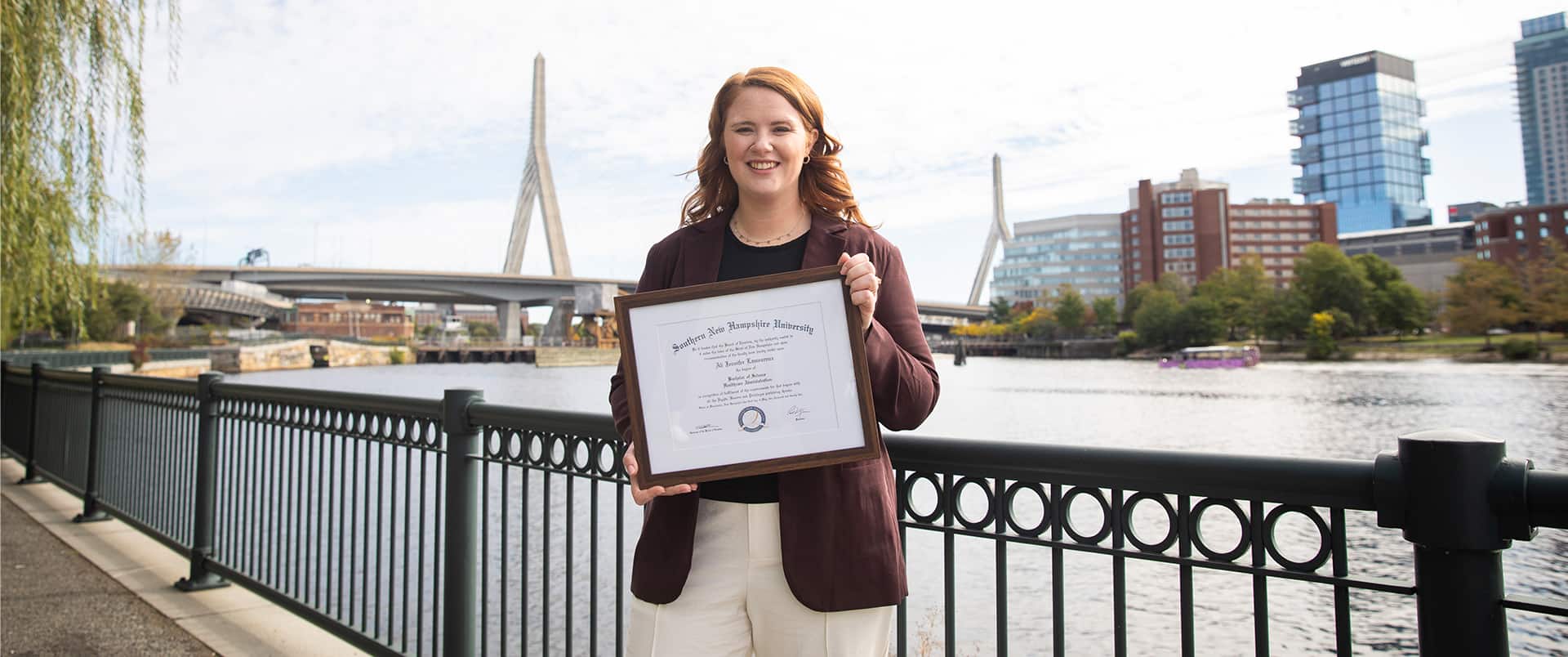 Ali Lamoreux, who earned her degree in 2022, holding her framed SNHU degree in front of a river with a tall bridge in the background.