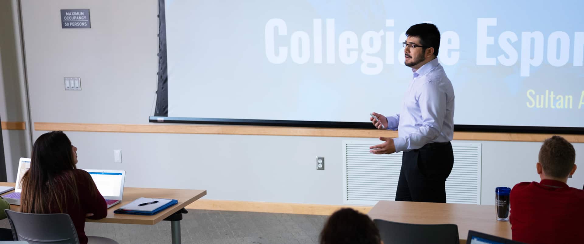Sultan Ahktar, who earned a degree from SNHU in 2019, standing in front of a college classroom with a projector screen behind him reading Collegiate Esports.