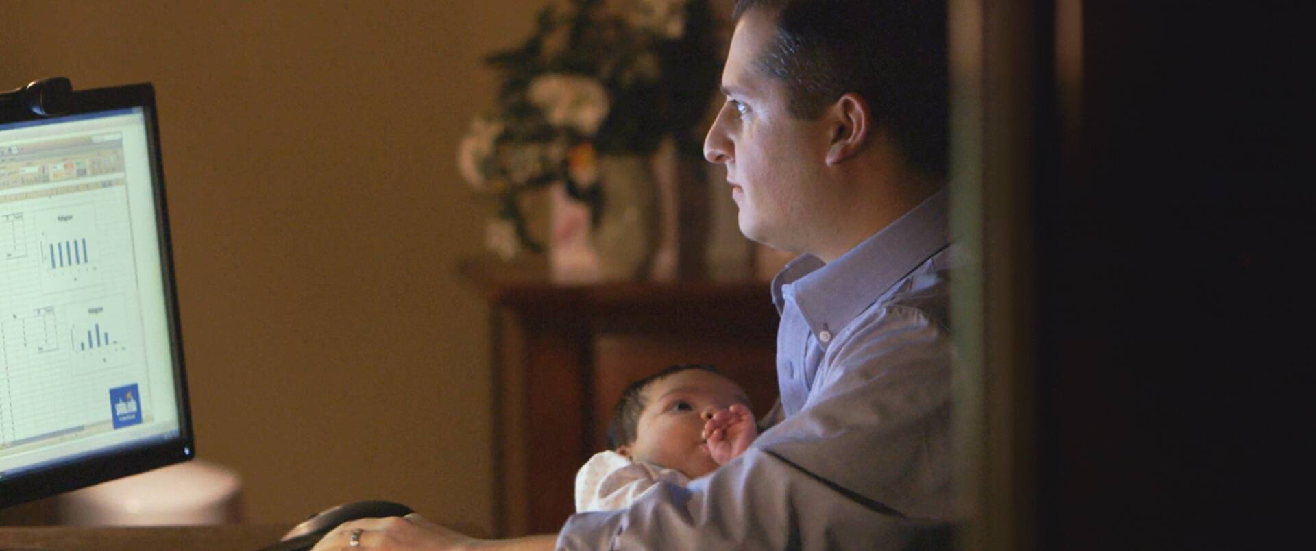 Peter Forcelli, a 2015 master's in healthcare administration alum, viewing charts on a computer while holding a baby.