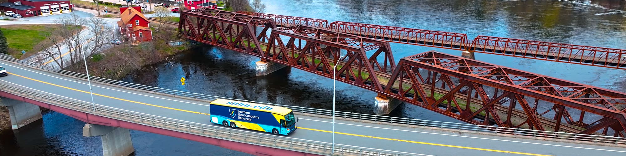 A yellow and blue coach bus with the words with SNHU’s logo on the side seen from above driving over a bridge.