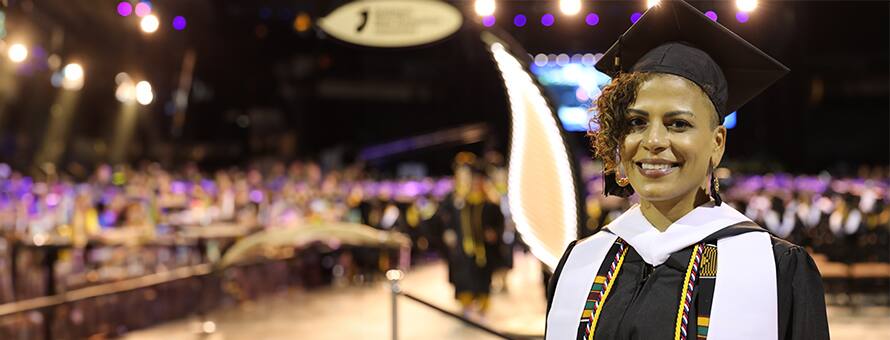 A woman wearing a black graduation cap and gown with a large crowd behind her inside an arena